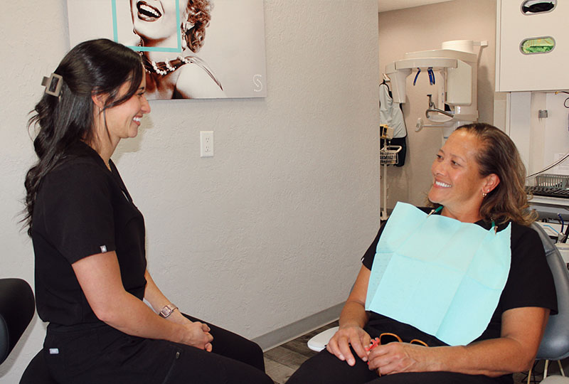 In the image, there are two women in a dental office setting. The woman on the right is sitting in a dental chair with her mouth open for treatment, while the woman standing next to her appears to be a dental professional, likely a dentist or hygienist, engaged in conversation with the seated patient.